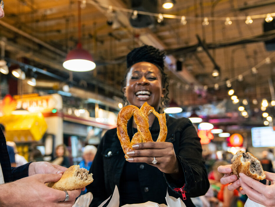 Three visitors share a laugh while enjoying Philadelphia food favorites, including a freshly baked soft pretzel, roast pork sandwich and cheesesteak inside Reading Terminal Market.