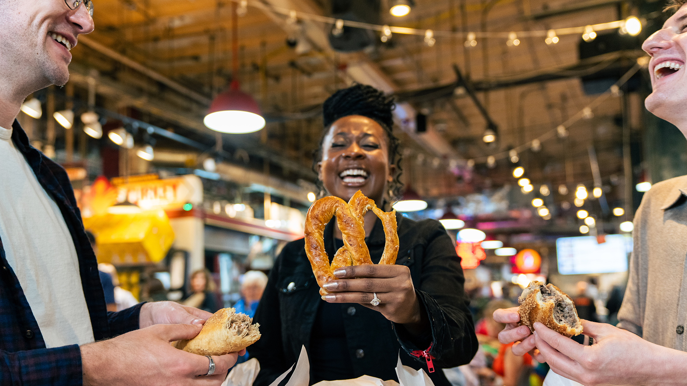 Three visitors share a laugh while enjoying Philadelphia food favorites, including a freshly baked soft pretzel, roast pork sandwich and cheesesteak inside Reading Terminal Market.