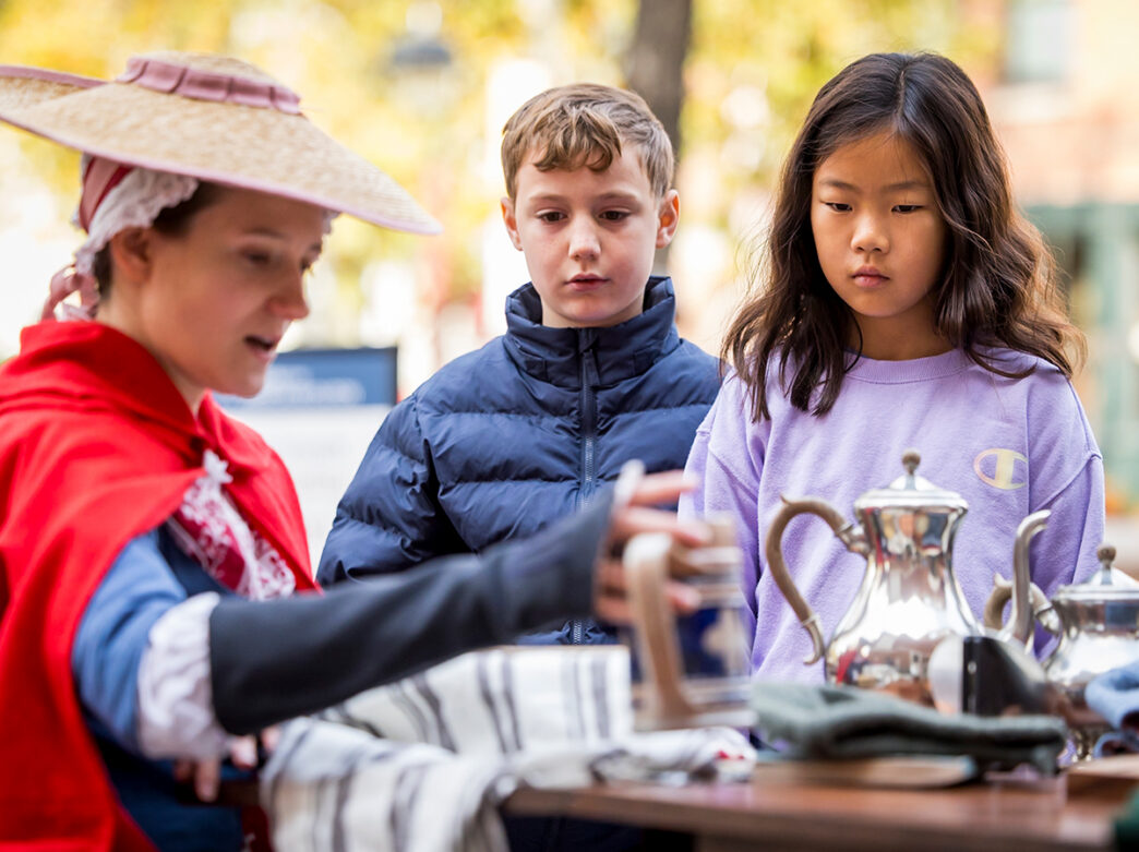 Two children watch a costumed historical interpreter demonstrate colonial-era objects and tableware during a Revolutionary Philadelphia program.