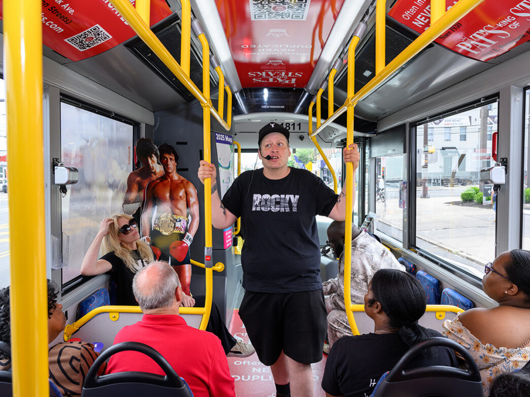 Un guía turístico con una camiseta negra de Rocky habla con los pasajeros sentados dentro de un autobús decorado con imágenes de Rocky, entre las que se incluye una figura recortada a tamaño real de un boxeador con un cinturón de campeón.