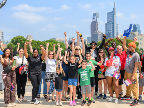 A large group of Rocky fans and tour participants raise their fists in a victory pose beside the Rocky statue, with the Philadelphia skyline visible behind them.