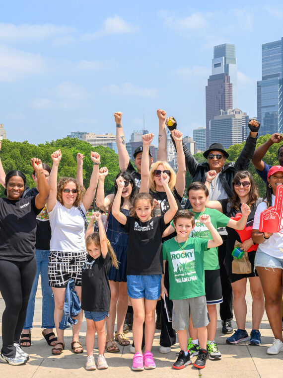 A large group of Rocky fans and tour participants raise their fists in a victory pose beside the Rocky statue, with the Philadelphia skyline visible behind them.
