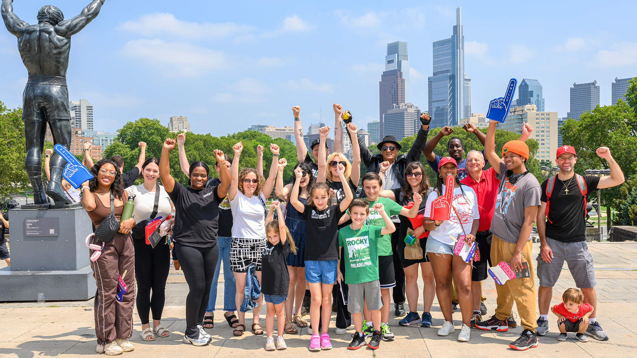 A large group of Rocky fans and tour participants raise their fists in a victory pose beside the Rocky statue, with the Philadelphia skyline visible behind them.