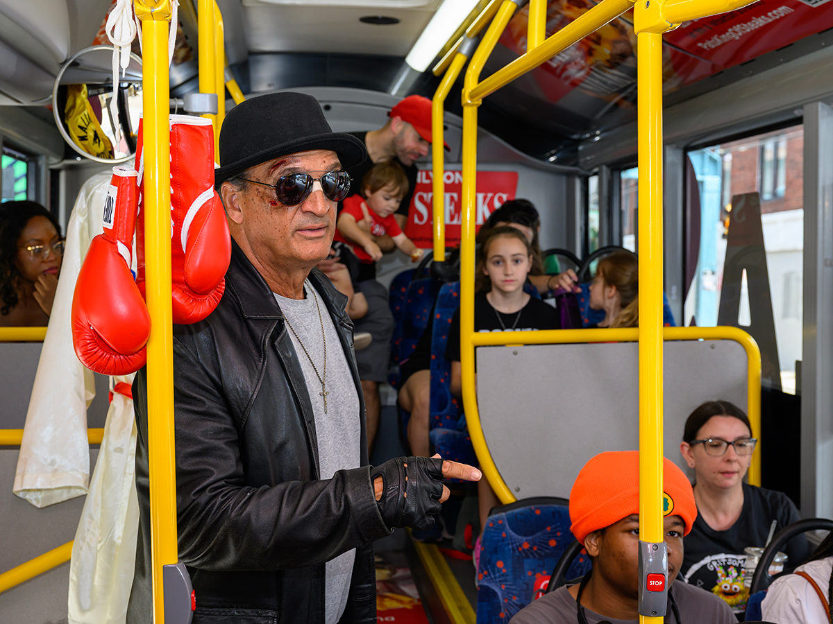 A rocky impersonator wearing a black hat, leather jacket and leather fingerless gloves stands inside a tour bus besides a pair of red boxing gloves hanging from above as passengers sit behind him.