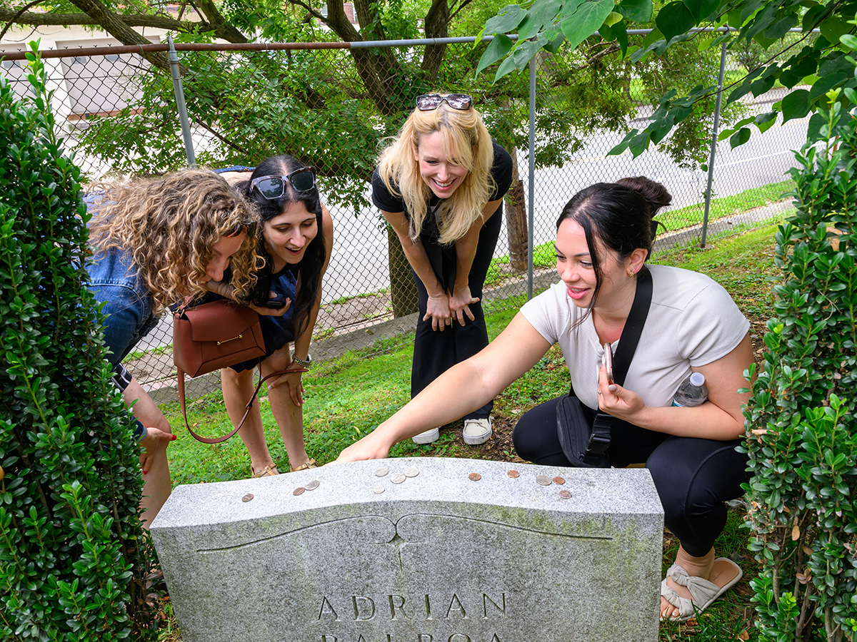 Four women gather around a headstone for Adrian Balboa at Laurel Hill Cemetery, with one pointing to the stone.