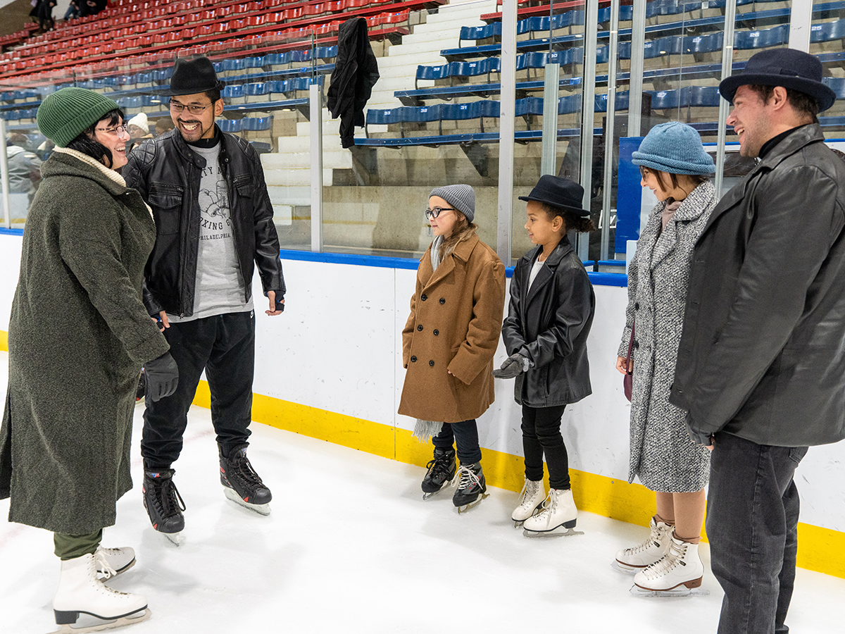 A group of adults and children dress in winter coats and hats smile together while wearing ice skates at an indoor ice rink.