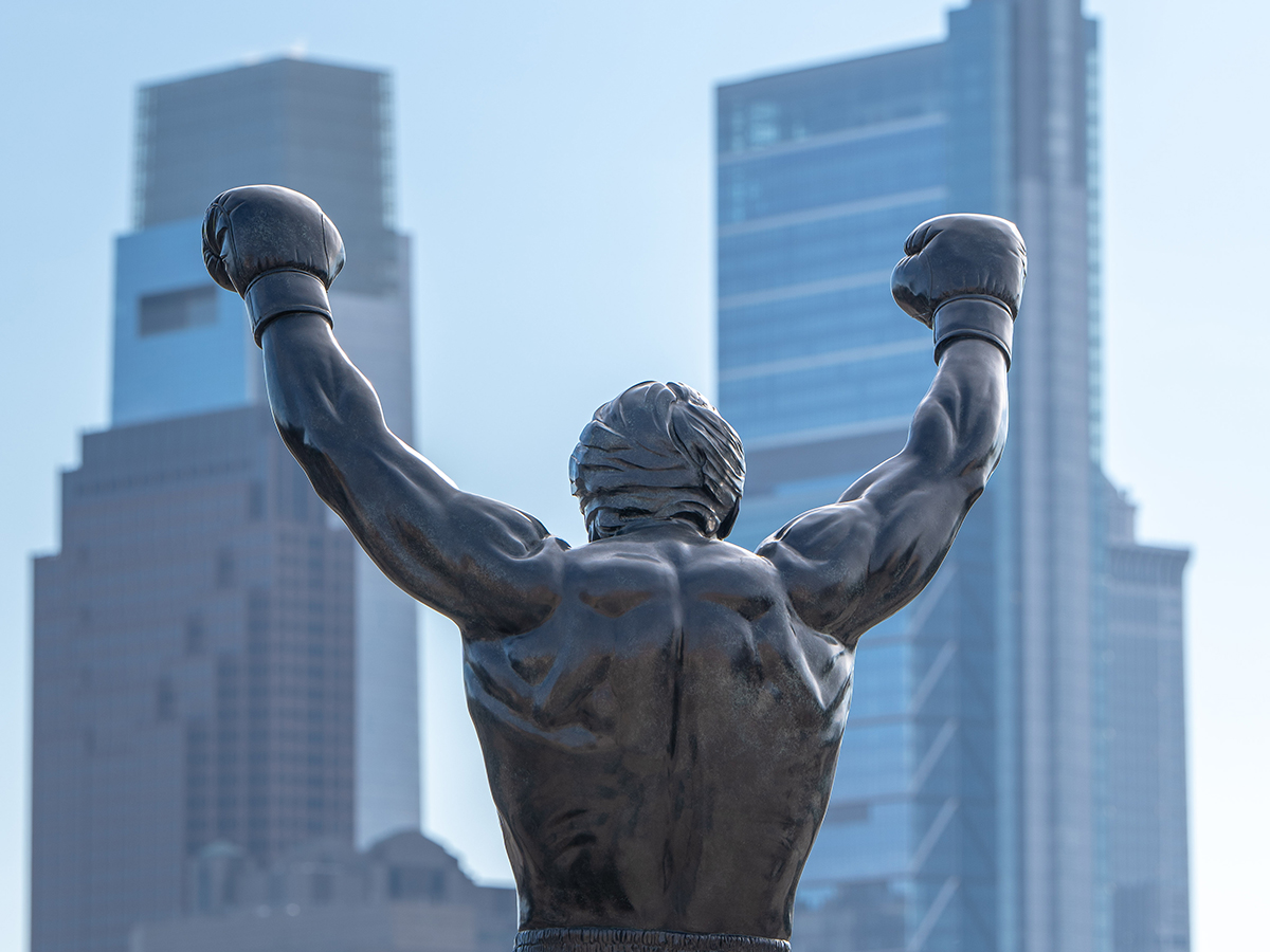 A bronze Rocky statue raises both boxing-gloved fists in victory, seen from behind with Philadelphia's skyscrapers in the background.