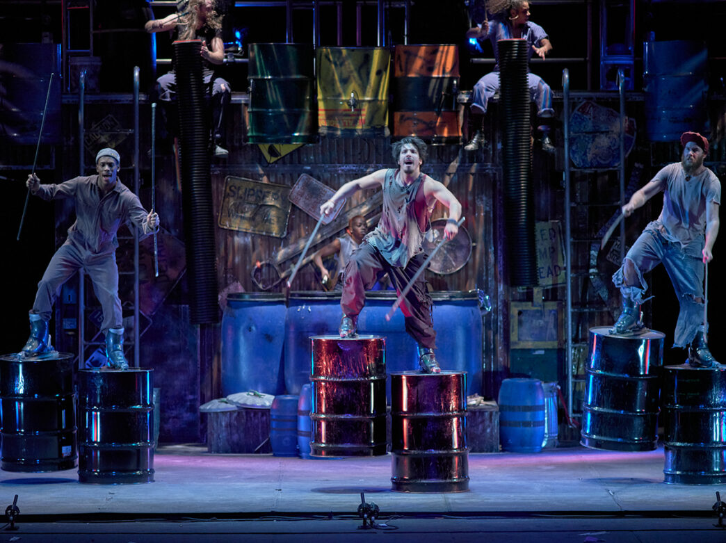 Performers in industrial style clothing use metal barrels and percussion sticks during an energetic performance of STOMP.