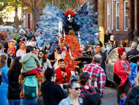 A lively Dia de los Muertos procession fills the streets of South Philadelphia, featuring participants in colorful traditional attire and elaborate skeleton makeup, plus a towering figure adorned with orange marigolds and dramatic blue wings stands in the center of the celebration.