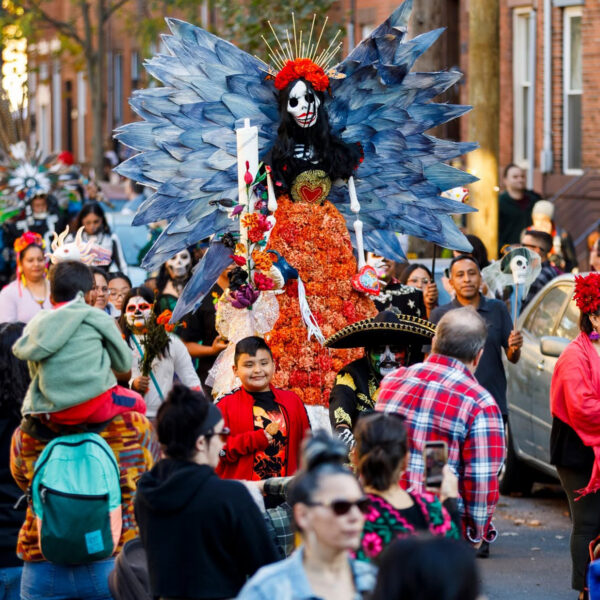 A lively Dia de los Muertos procession fills the streets of South Philadelphia, featuring participants in colorful traditional attire and elaborate skeleton makeup, plus a towering figure adorned with orange marigolds and dramatic blue wings stands in the center of the celebration.