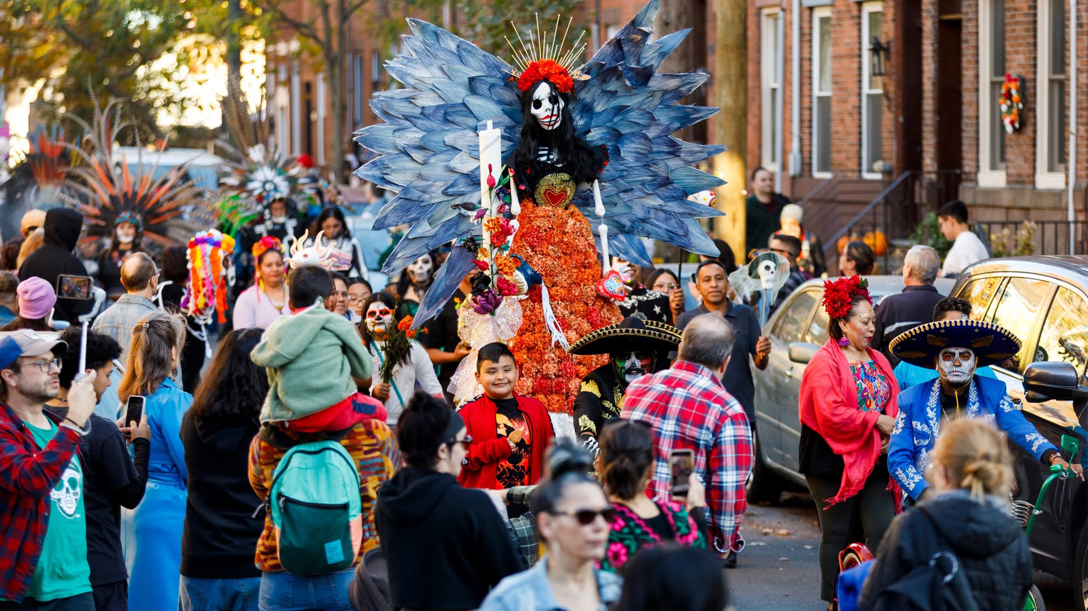 Una animada procesión del Día de los Muertos llena las calles del sur de Filadelfia, con participantes vestidos con coloridos trajes tradicionales y elaborados maquillajes de esqueletos; además, una imponente figura adornada con caléndulas anaranjadas y espectaculares alas azules se erige en el centro de la celebración.