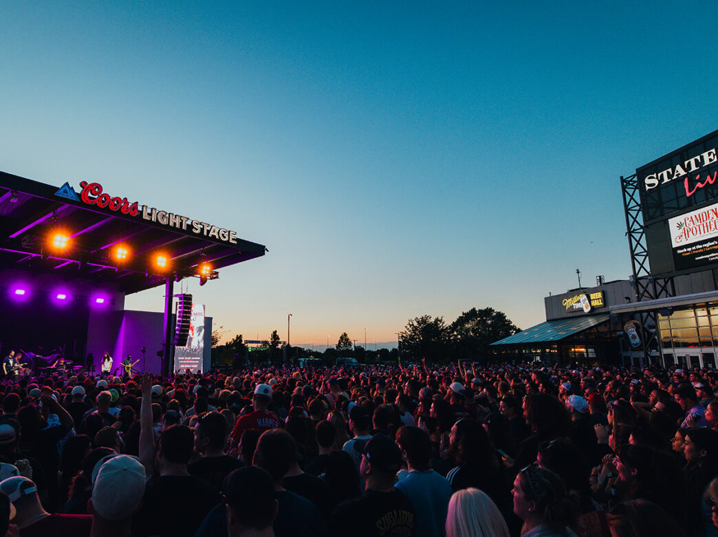 Una gran multitud se reúne para disfrutar de un concierto en vivo en un escenario al aire libre en Stateside Live!, con una colorida iluminación escénica que ilumina el cielo nocturno.