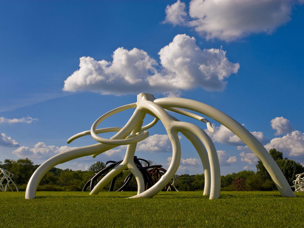A large, abstract white steel sculpture by artist Steve Tobin, featuring twisting and interconnected root-like forms, stands in a grassy field beneath a bright blue sky with fluffy clouds.