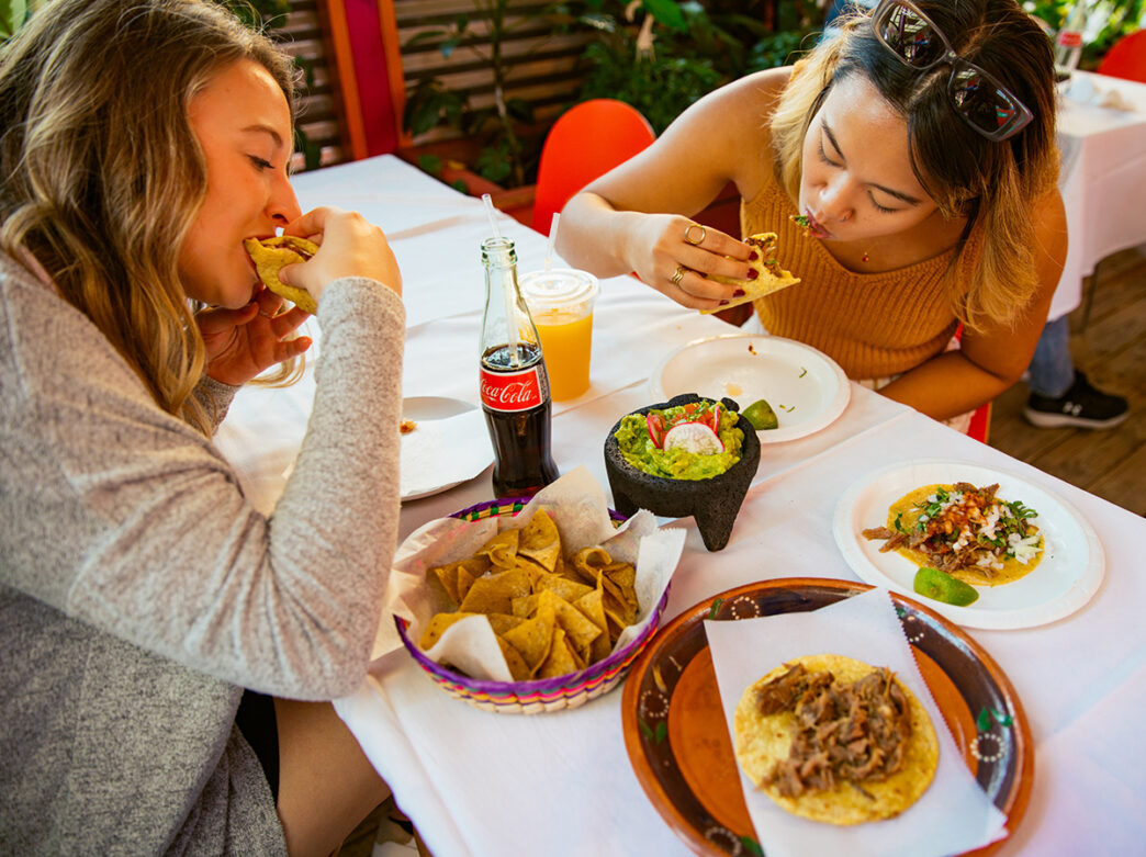 Two women enjoy tacos at South Philly Barbacoa, while seated at a table with a white tablecloth, chips, guacamole, soda and more.