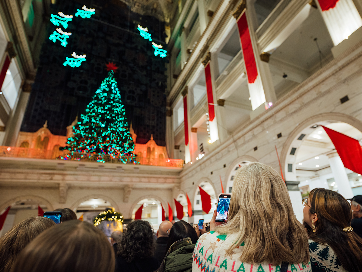 Visitors gather inside a historic building to enjoy the annual holiday light show, featuring a towering glowing Christmas tree, surrounded by illuminated reindeer and festive decorations.