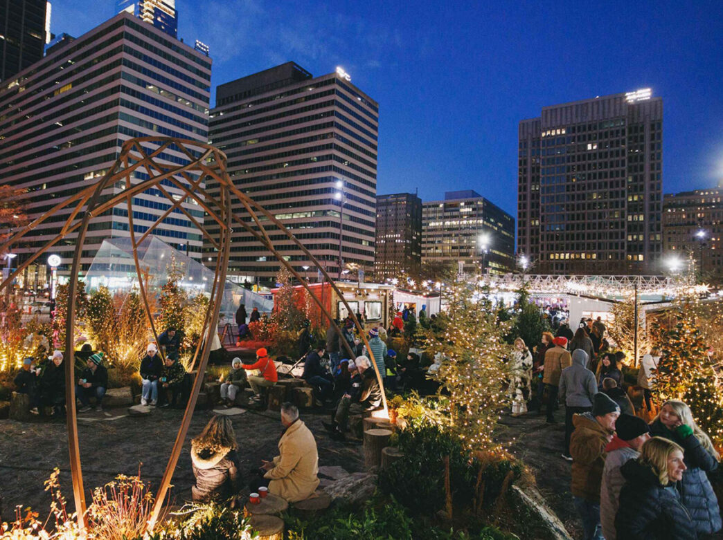 Visitors gather among illuminated Christmas trees and festive greenery at Wintergarden in Dilworth Park, with Center City buildings rising against the evening sky.