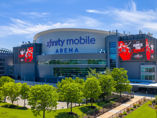 The exterior of Xfinity Mobile Arena on a sunny day, with large Philadelphia Flyers logos displayed on digital screens above an entrance to the arena.