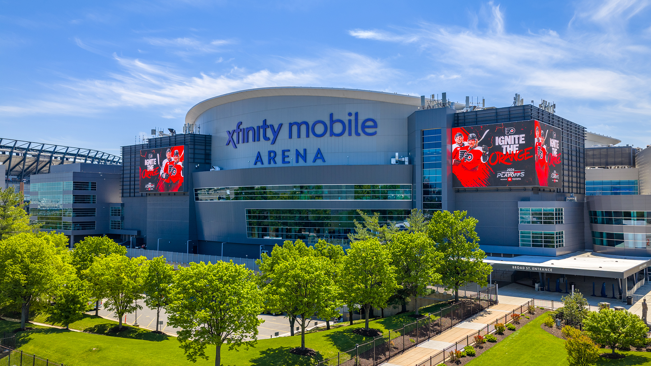 The exterior of Xfinity Mobile Arena on a sunny day, with large Philadelphia Flyers logos displayed on digital screens above an entrance to the arena.