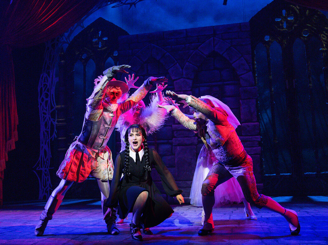 Performers dressed in elaborate costumes surround Wednesday Addams dressed in all black with braided pigtails, during a scene from The Addams Family, illuminated by dramatic blue stage lighting.