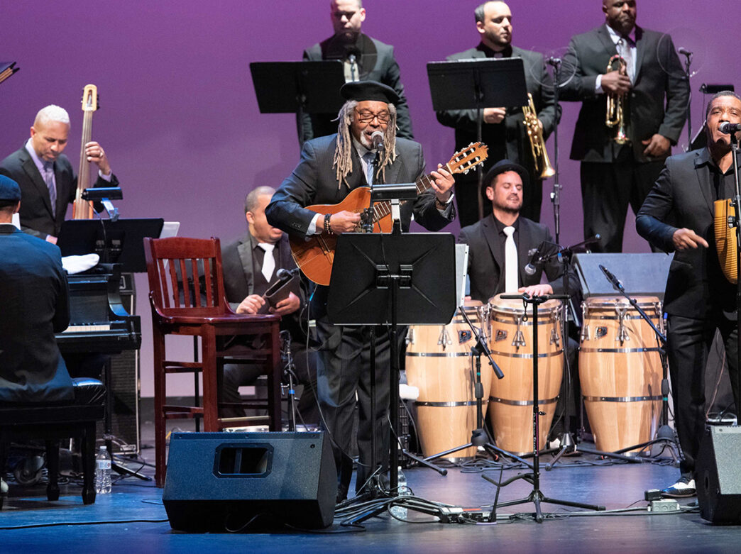 The Afro-Cuban All Stars perform on stage, featuring a lead vocalist playing guitar alongside musicians on piano, upright bass, percussion and brass instruments against a purple stage background.
