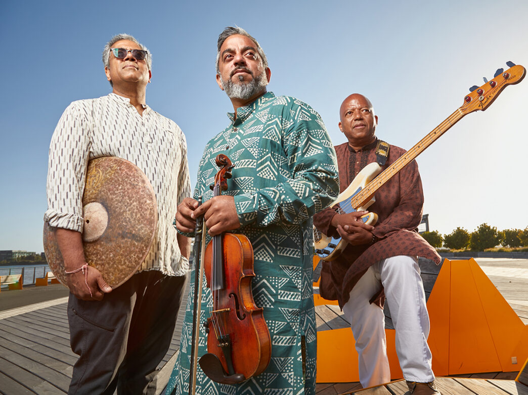 Three members of the Arun Ramamurthy Trio pose outdoors along a waterfront , holding a violin, a bass guitar and a percussion instrument.