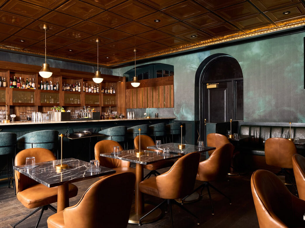An elegant cocktail bar featuring tan leather chairs, black marble tables, teal velvet barstools, wood-paneled shelves and warm pendant lighting.