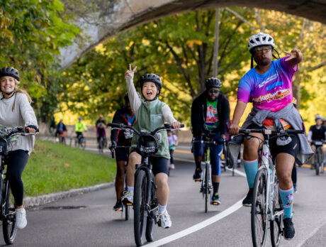 A group of cyclists smile and flash peace signs while riding beneath a stone bridge along a tree-lined road during the Philly Bike Ride.