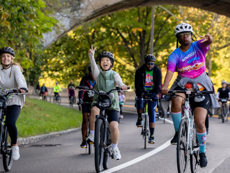 A group of cyclists smile and flash peace signs while riding beneath a stone bridge along a tree-lined road during the Philly Bike Ride.