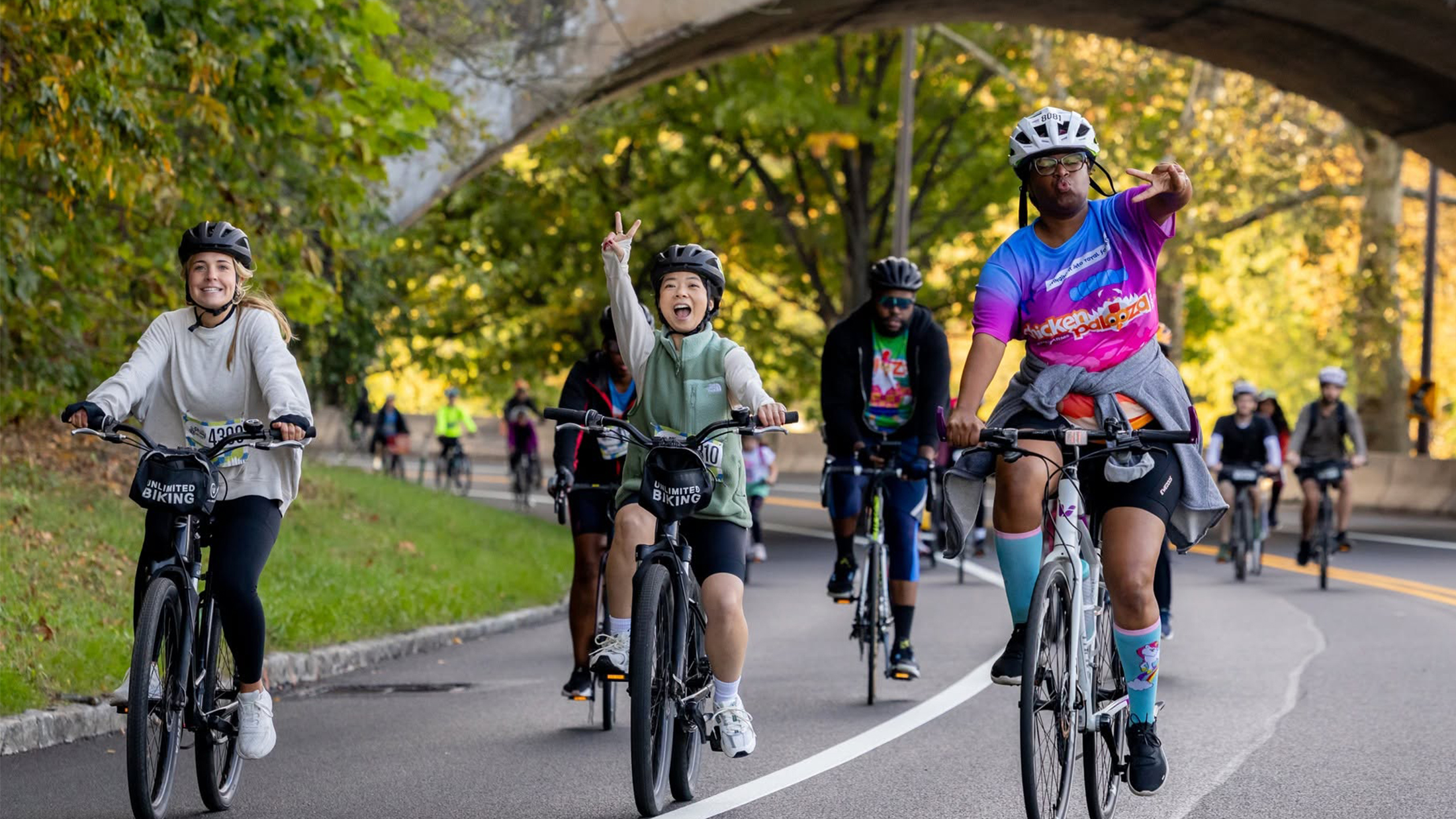 A group of cyclists smile and flash peace signs while riding beneath a stone bridge along a tree-lined road during the Philly Bike Ride.