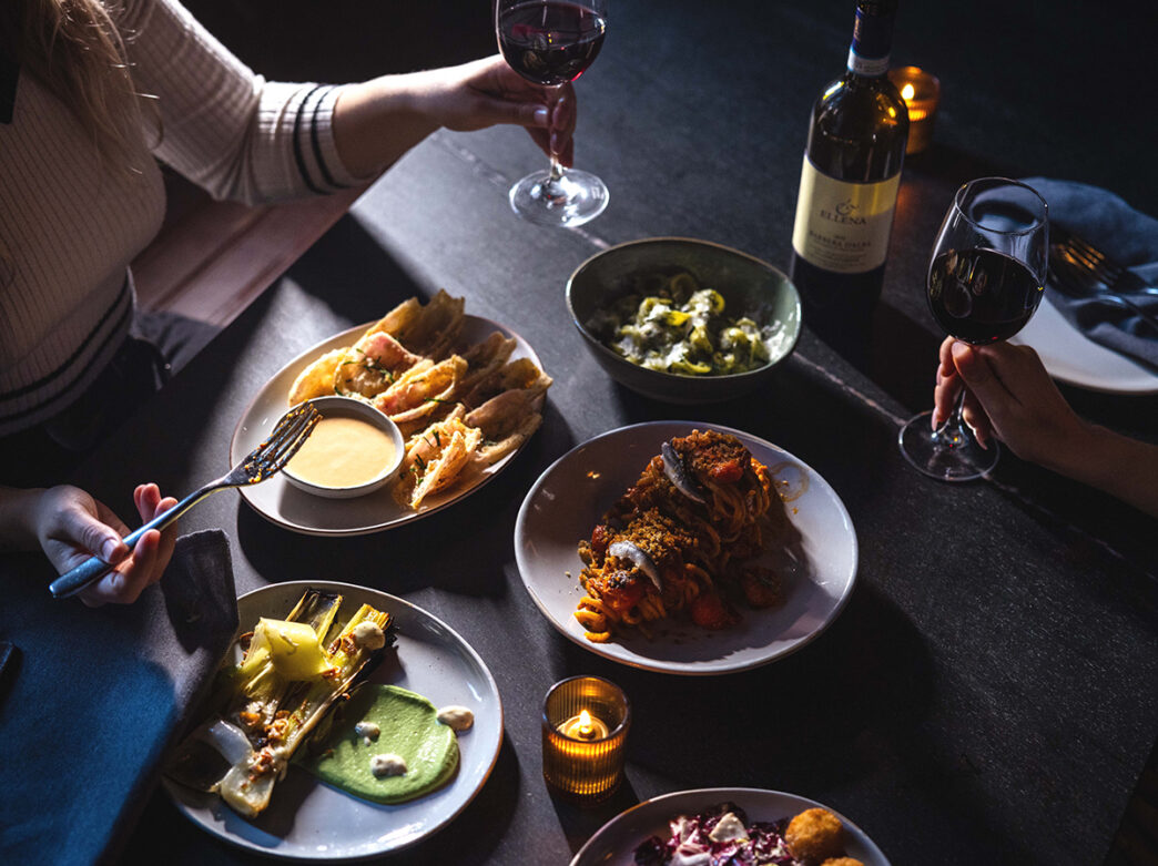 Two diners enjoy glasses of red wine alongside a spread of small plates, including pasta, roasted vegetables and other dishes.