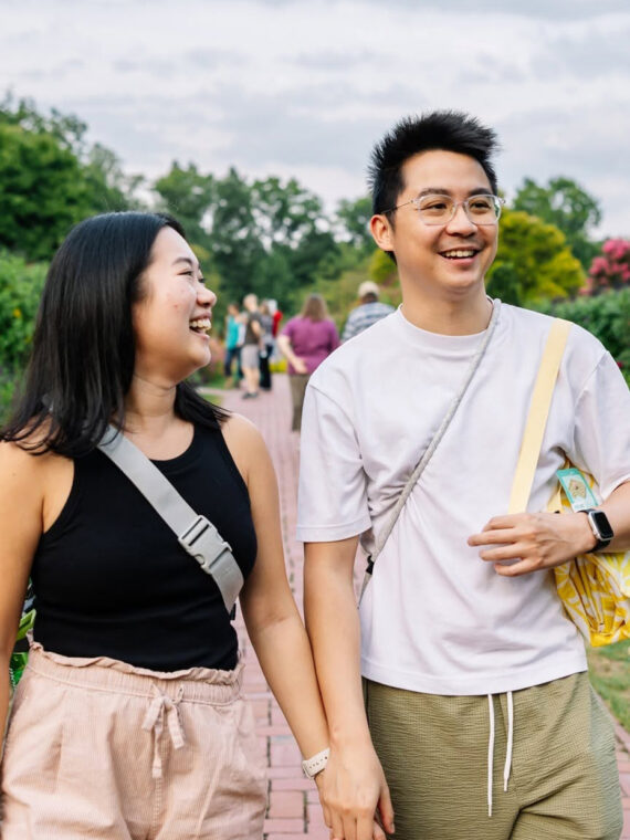 Una pareja sonriente pasea de la mano por un sendero de ladrillo en Longwood Gardens, rodeada de flores coloridas, exuberante vegetación y vibrantes flores de temporada.