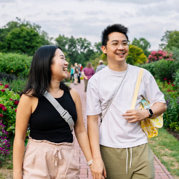 A smiling couple strolls hand in hand along a brick pathway at Longwood Gardens, surrounded by colorful flowers, lush greenery and vibrant seasonal blooms.