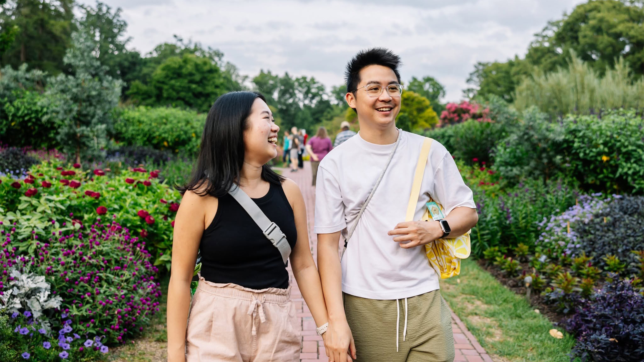 A smiling couple strolls hand in hand along a brick pathway at Longwood Gardens, surrounded by colorful flowers, lush greenery and vibrant seasonal blooms.