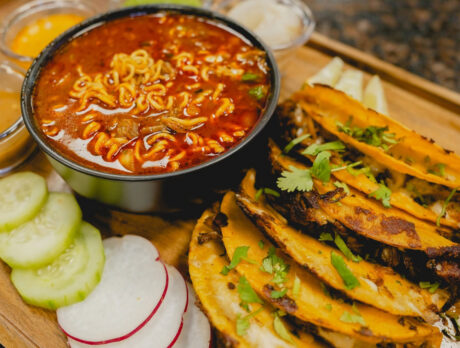 A spread of crispy, golden-brown birria tacos topped with fresh cilantro, served alongside a bowl of rich, red broth with noodles for dipping.