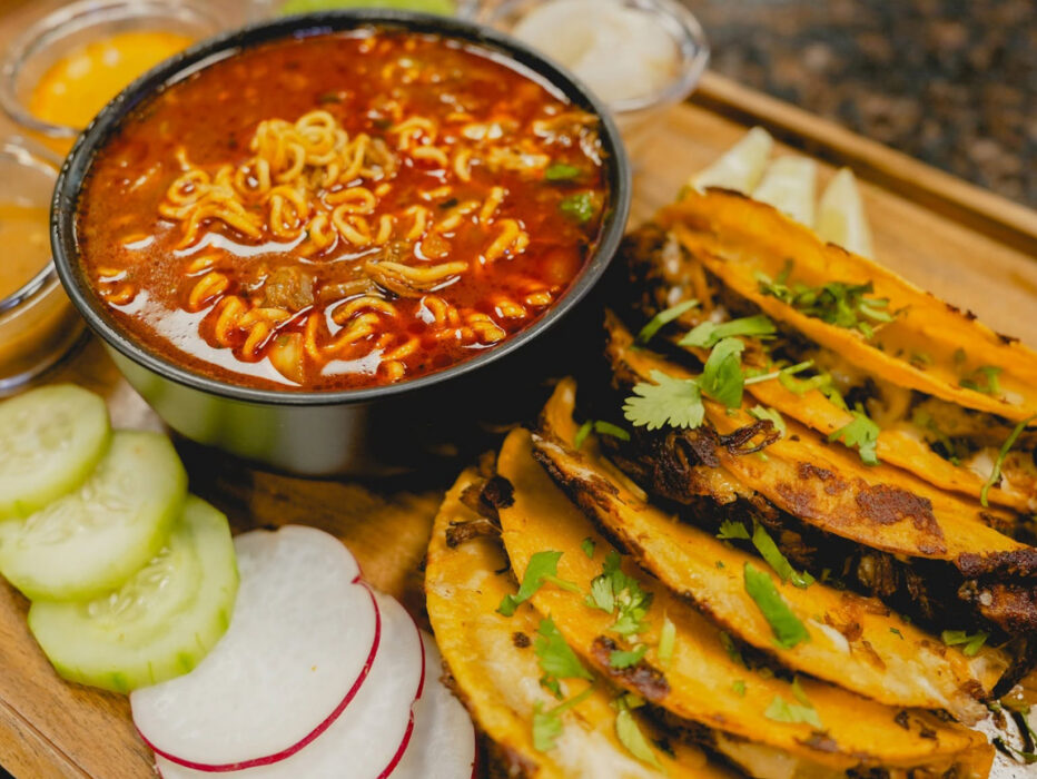 A spread of crispy, golden-brown birria tacos topped with fresh cilantro, served alongside a bowl of rich, red broth with noodles for dipping.