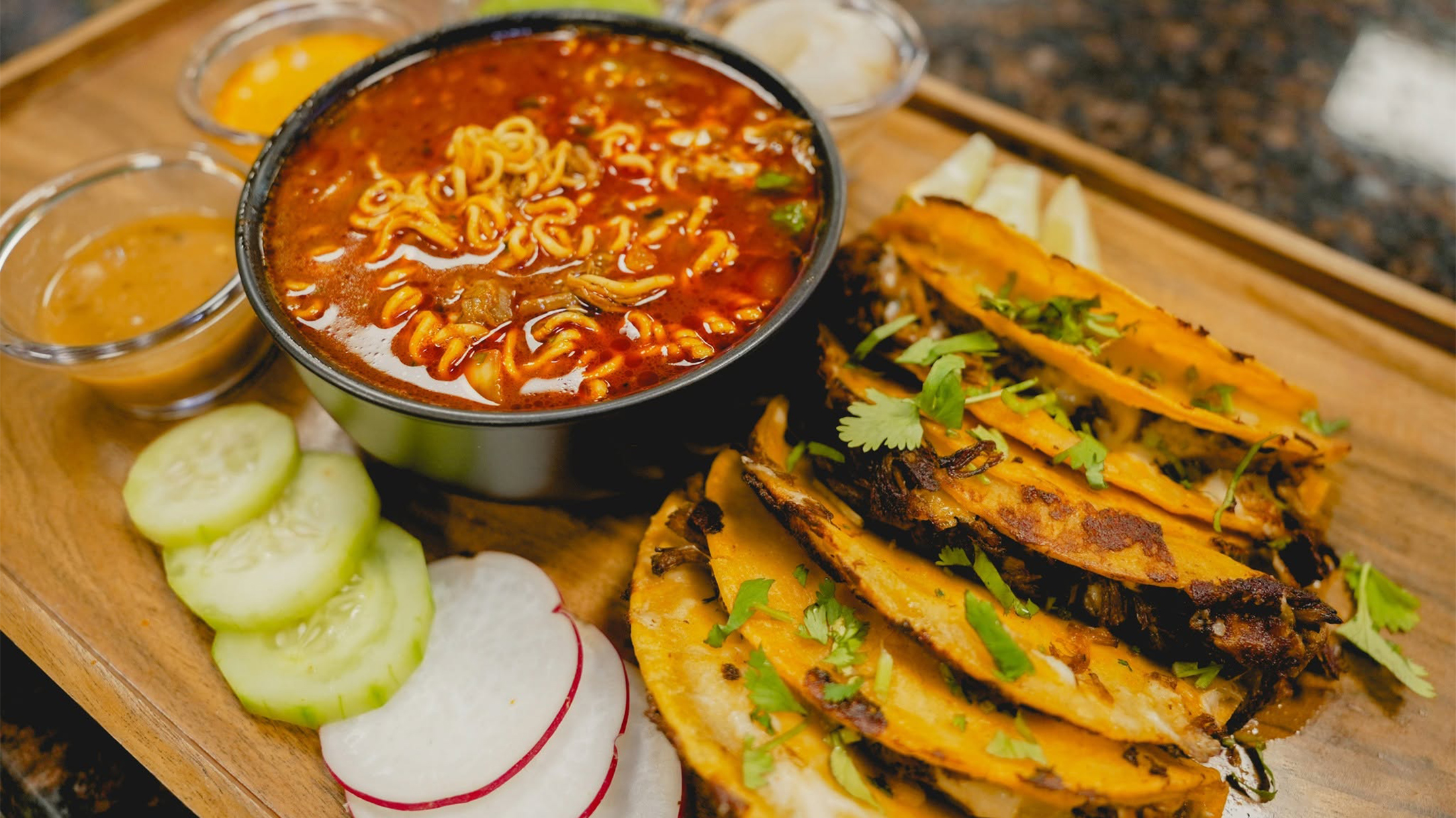 Una variedad de tacos de birria crujientes y dorados, adornados con cilantro fresco, servidos junto con un tazón de caldo rojo y sustancioso con fideos para mojar.