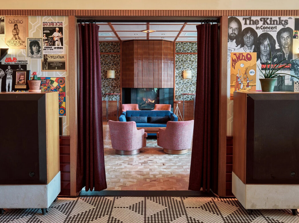 A retro-inspired lounge featuring walls covered in vintage concert posters, large speakers, patterned tile floors and a cozy seating area with pink chairs, a blue sofa and fireplace.