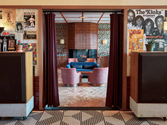 A retro-inspired lounge featuring walls covered in vintage concert posters, large speakers, patterned tile floors and a cozy seating area with pink chairs, a blue sofa and fireplace.