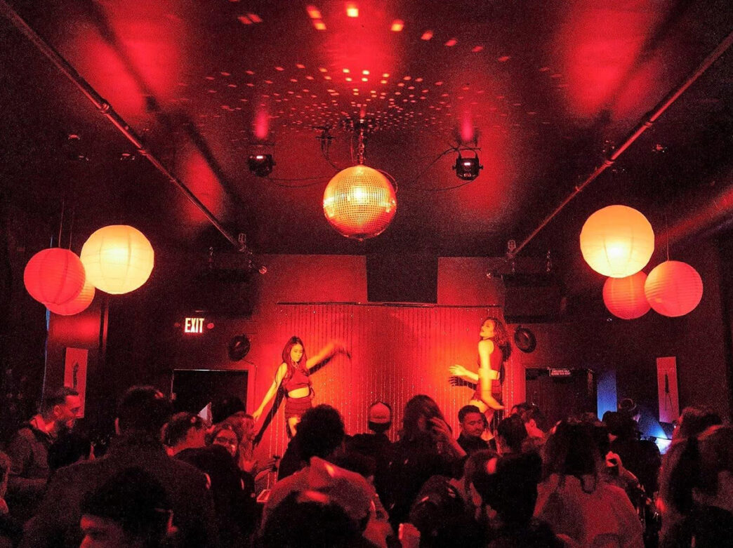 A crowded dance floor glows under vibrant red lighting, with two dancers performing on stage beneath a disco ball and hanging paper lanterns.