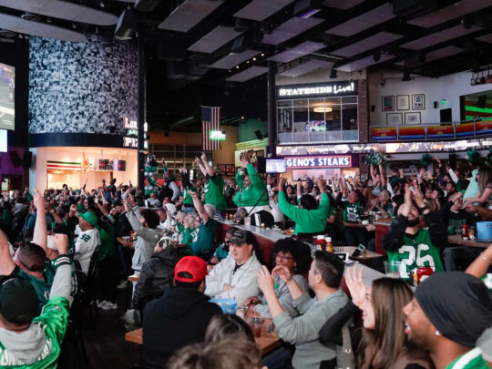 A packed crowd of Philadelphia Eagles fans cheer with their arms raised while watching a football game on a giant screen.