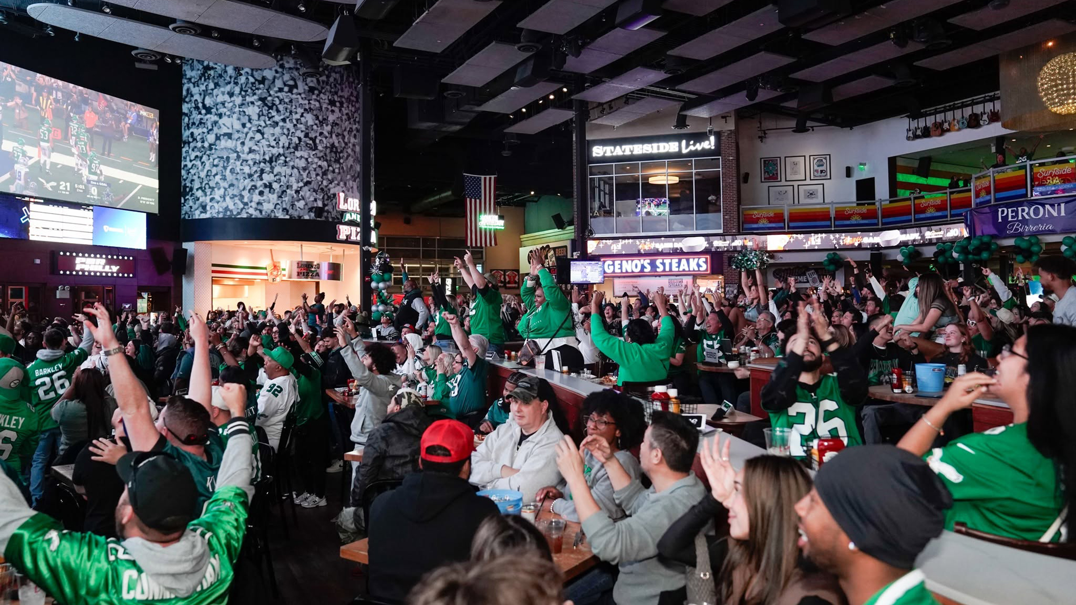 A packed crowd of Philadelphia Eagles fans cheer with their arms raised while watching a football game on a giant screen.