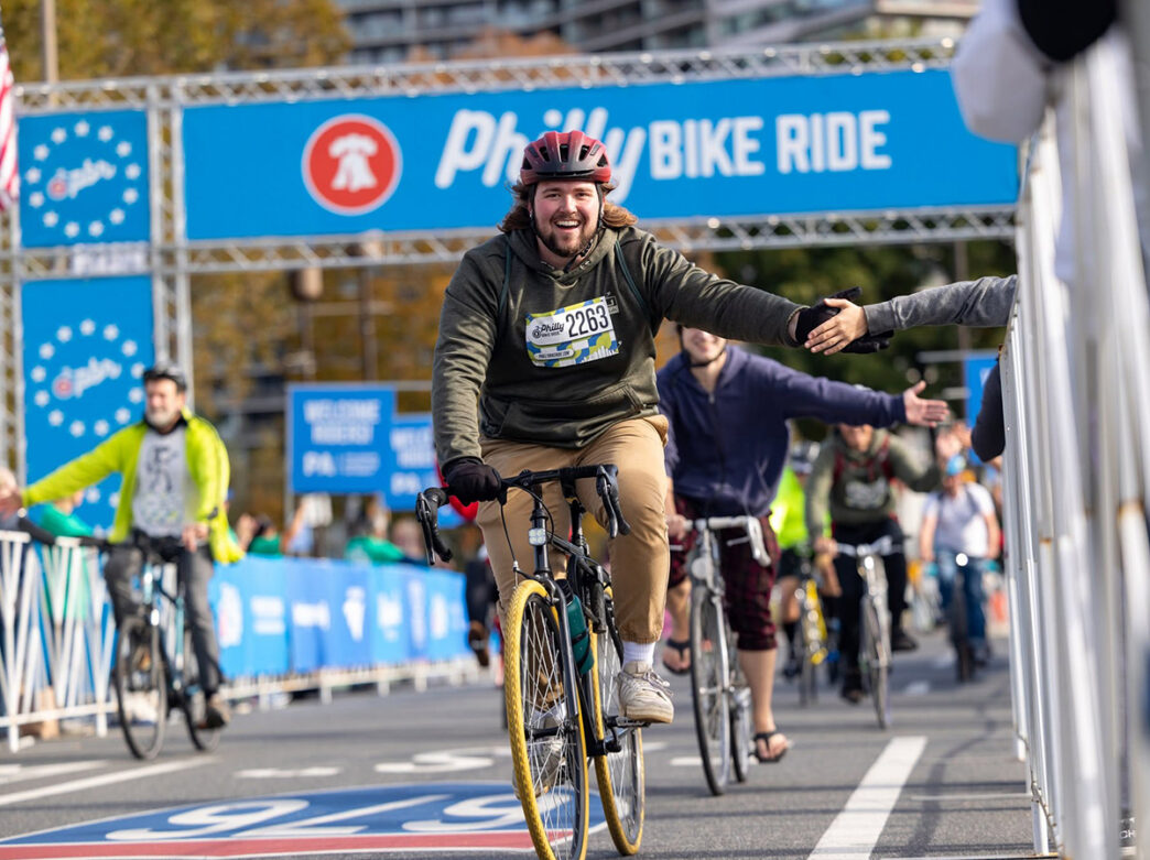 A smiling cyclist wearing a red helmet reached out for a high-five while riding through the finish line of the Philly Bike Ride, with fellow participants cycling behind them.