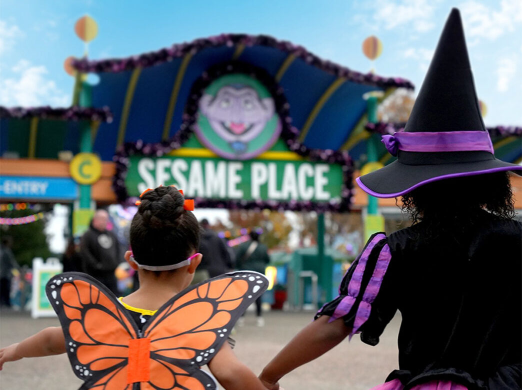 Two children wearing Halloween costumes, one wearing a pair of orange butterfly wings and the other in a purple-and-black witch outfit, walk hand-in-hand toward the decorated entrance of Sesame Place.