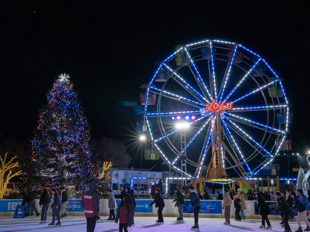 Ice skaters enjoy a festive evening beneath a brightly illuminated Ferris wheel and colorful Christmas tree at an outdoor ice skating rink.
