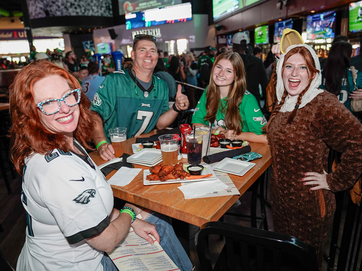 Philadelphia Eagles fans wearing team jerseys pose around a table of wings and beer at Stateside Live!, alongside a fan dressed in an eagle costume.