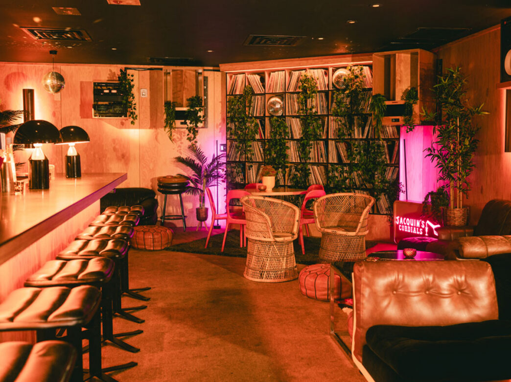 A cozy retro-inspired lounge featuring leather seating, wicker chairs, shelves of vinyl records, lush greenery and pink lighting.