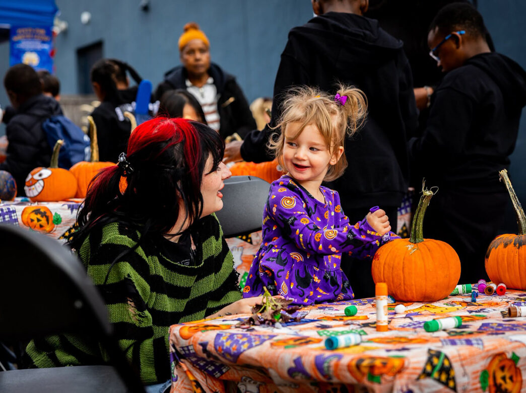 A young girl in a purple Halloween dress decorates a pumpkin alongside an adult at a festive craft table during Manayunk Fall Fest.