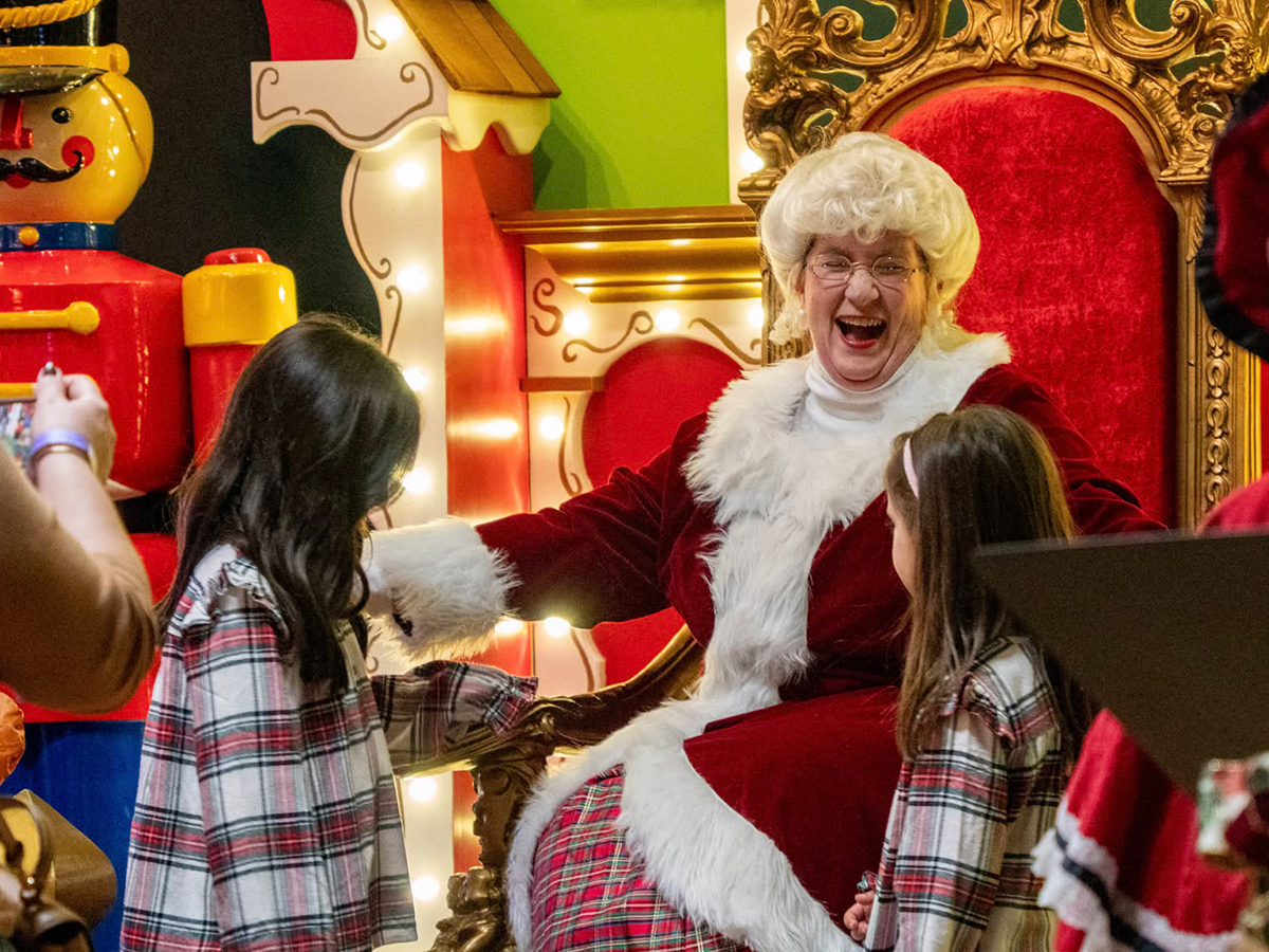 Mrs. Claus smiles and greets two young visitors wearing red plaid pajamas while seated on an ornate velvet red-and-gold throne surrounded by festive holiday decorations.