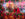 Two television hosts smile behind a vibrant display of pink and orange flowers during Philadelphia's 6abc Dunkin' Thanksgiving Day Parade, with colorful holiday decorations and parade festivities in the background.