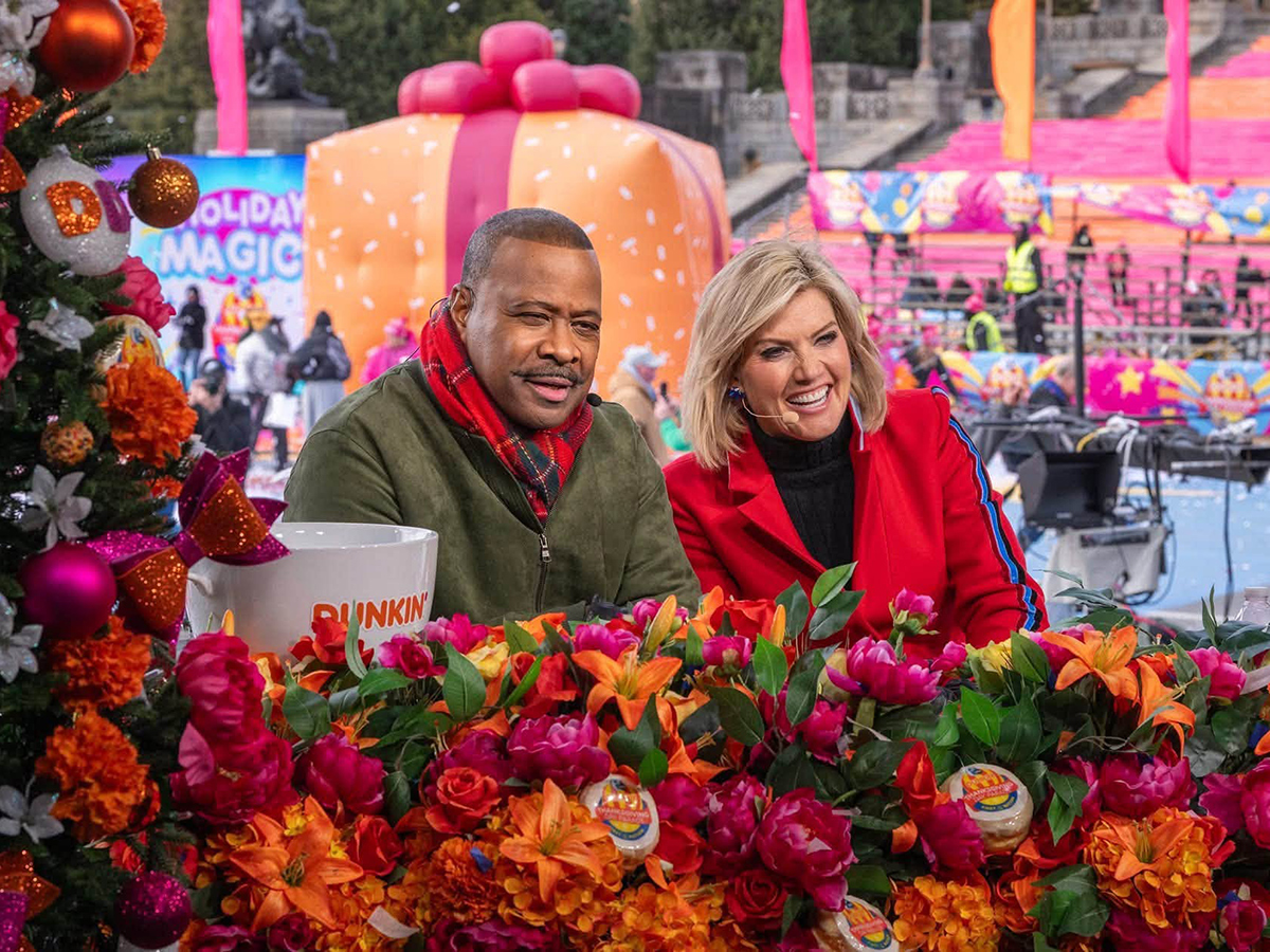 Two television hosts smile behind a vibrant display of pink and orange flowers during Philadelphia's 6abc Dunkin' Thanksgiving Day Parade, with colorful holiday decorations and parade festivities in the background.